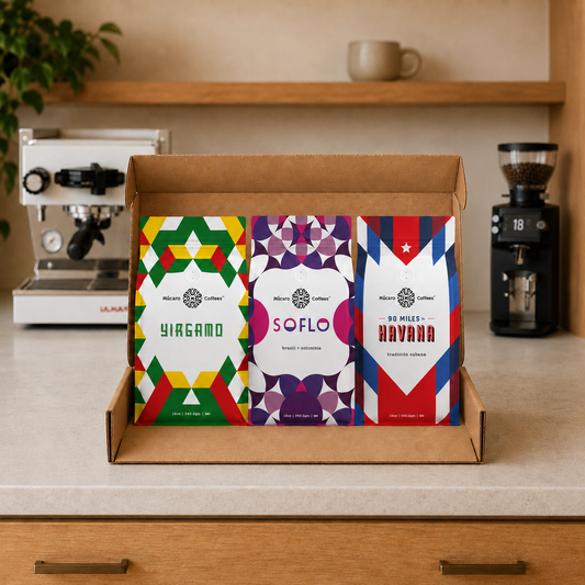 A cardboard box containing three bags of Mucaro Coffee beans (Yirgamo, Soflo, and 90 Miles to Havana) arranged on a kitchen counter with a coffee grinder and espresso machine in the background.
