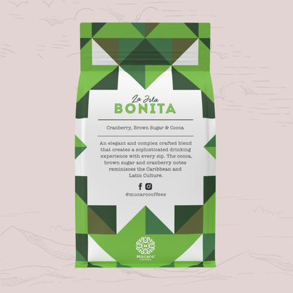 A Mucaro Coffees stand-up coffee bag with a green and white geometric pattern, featuring the brand name 'La Isla BONITA' on the front label. The bag is centered against a pale pink background with faint hand-drawn mountain silhouettes.