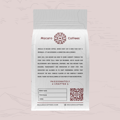 A white bag of Mucaro Coffee's 'Passionately Crafted' coffee stands against a light pink background with faint line drawings of mountains and clouds. The bag features a gray top seal, black descriptive text, and dark red branding and logo details.