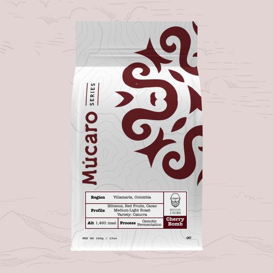 A stand-up pouch of Mucaro Series Cherry Bomb coffee beans, featuring a white bag with dark red tribal patterns and text, set against a light pink background with faint mountain silhouettes.
