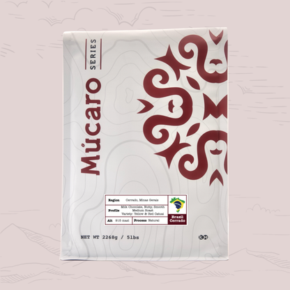 A white bag of Mucaro Series coffee from Cerrado Mineiro, Brazil featuring a prominent dark red graphic pattern on the right side and product details at the bottom, set against a light pink background with faint mountain silhouettes.