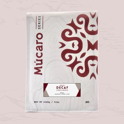 A package of Mucaro Series Decaf Single Origin coffee from Mexico on a light pink background with subtle mountain line drawings. The package is white with dark red text and a large red decorative pattern on the right side.