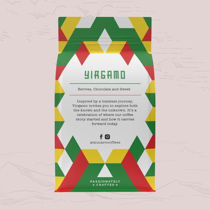 A bag of Mucaro Coffee's 'Yiragamo' coffee blend stands upright against a light pink background with faint hand-drawn mountain outlines. The packaging features a colorful geometric pattern of red, green, and yellow triangles framing a white center panel with product details.