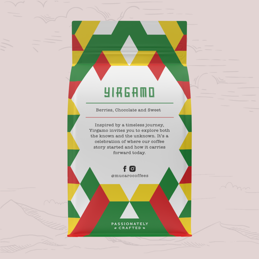 A bag of Mucaro Coffee's 'Yiragamo' coffee blend stands upright against a light pink background with faint hand-drawn mountain outlines. The packaging features a colorful geometric pattern of red, green, and yellow triangles framing a white center panel with product details.
