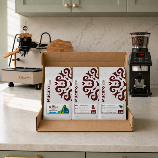 Three Mucaro Single Origins coffee bags displayed in an open cardboard box on a kitchen countertop, with a coffee machine and grinder in the background.